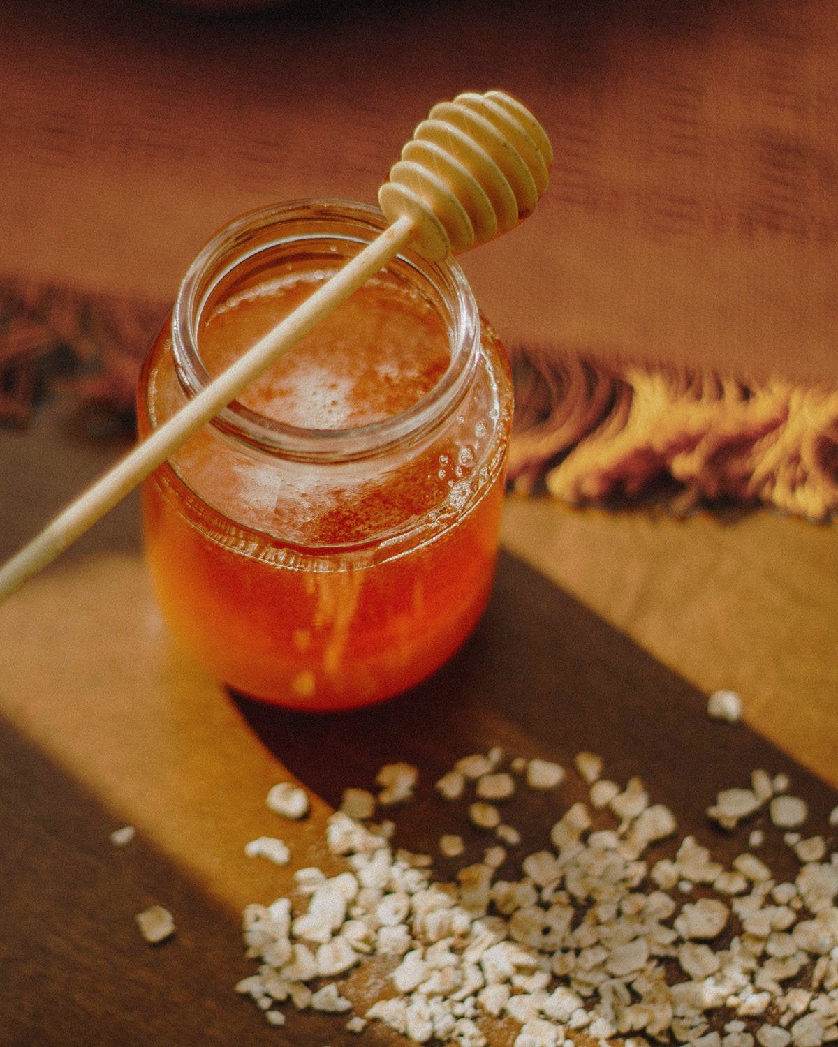 Honey jar with wooden dipper and scattered oats on a warm wooden cutting board — natural light still life