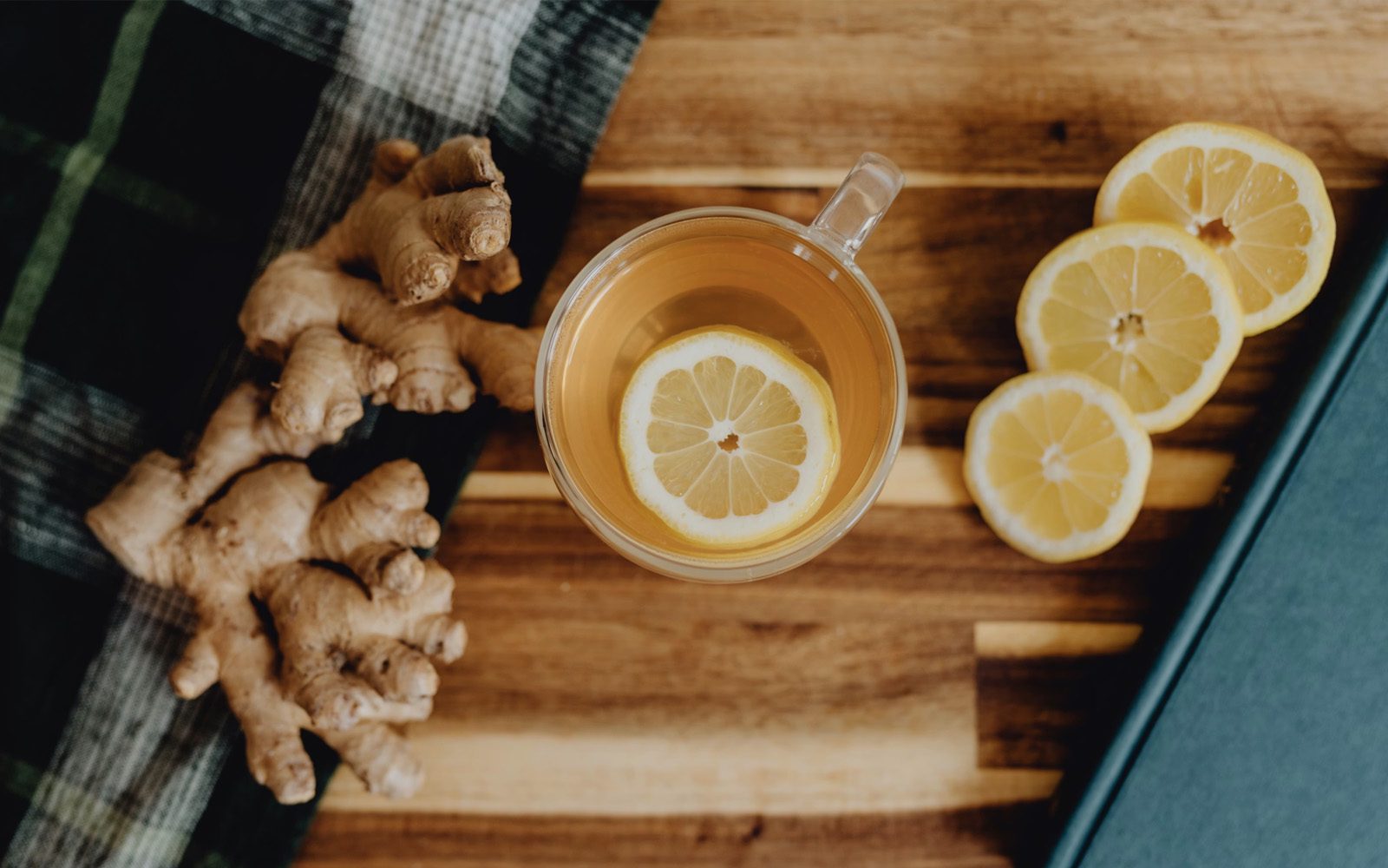 Ginger root, fresh lemon slices, and a glass cup of tea with a lemon round, arranged overhead on a warm wooden cutting board