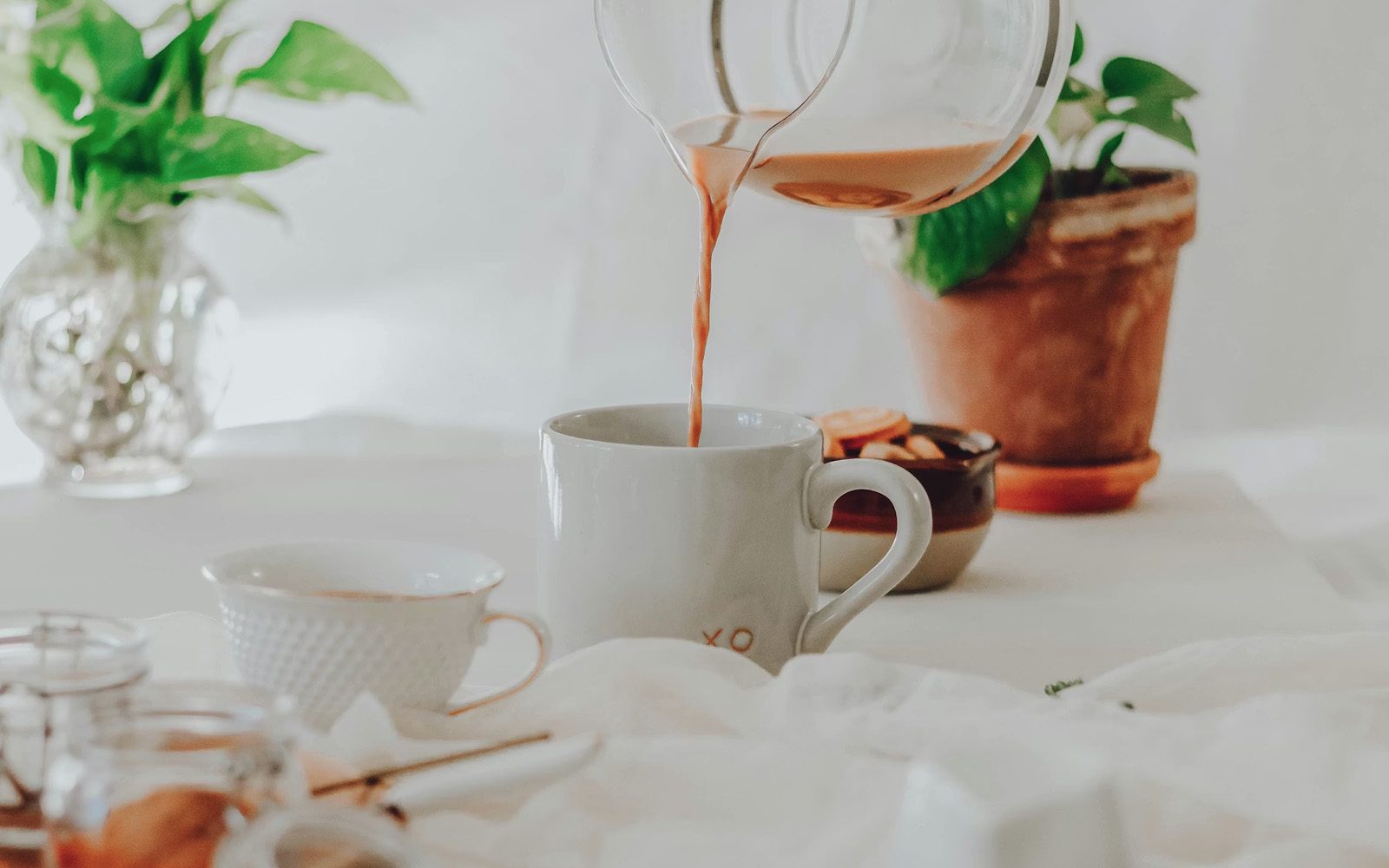 A glass pitcher pours tea into a white ceramic mug at a sunlit table, with potted plants and linen in soft morning light